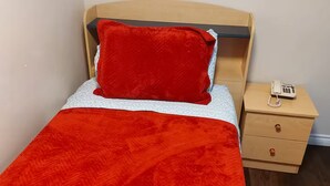 Un lit simple avec une couverture et un oreiller rouges, une tête de lit en bois, une table de chevet avec un téléphone et un petit tiroir.