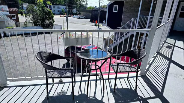 Un balcon avec deux chaises et une petite table, donnant sur une rue bordée de voitures garées et d’immeubles.