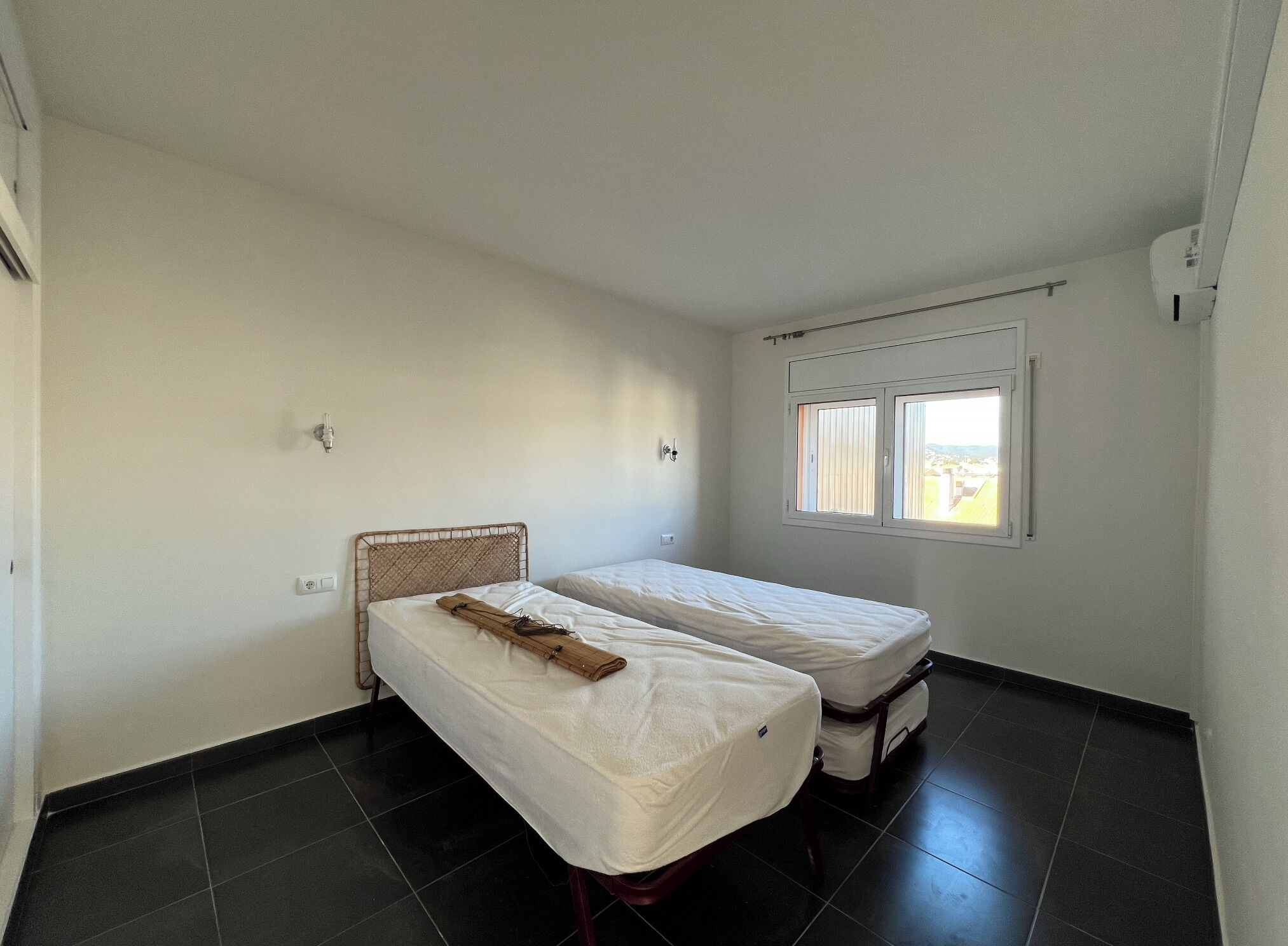 Una habitación pequeña y limpia con dos camas individuales, una ventana con persianas y una pared blanca sencilla.