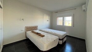Ein kleiner, sauberer Raum mit zwei Einzelbetten, einem Fenster mit Jalousien und einer schlichten weißen Wand.