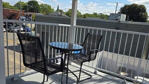 Un espace extérieur abrité, équipé d’une table et de deux chaises, d’une rambarde blanche, et offrant une vue sur un parking et des bâtiments en arrière-plan.