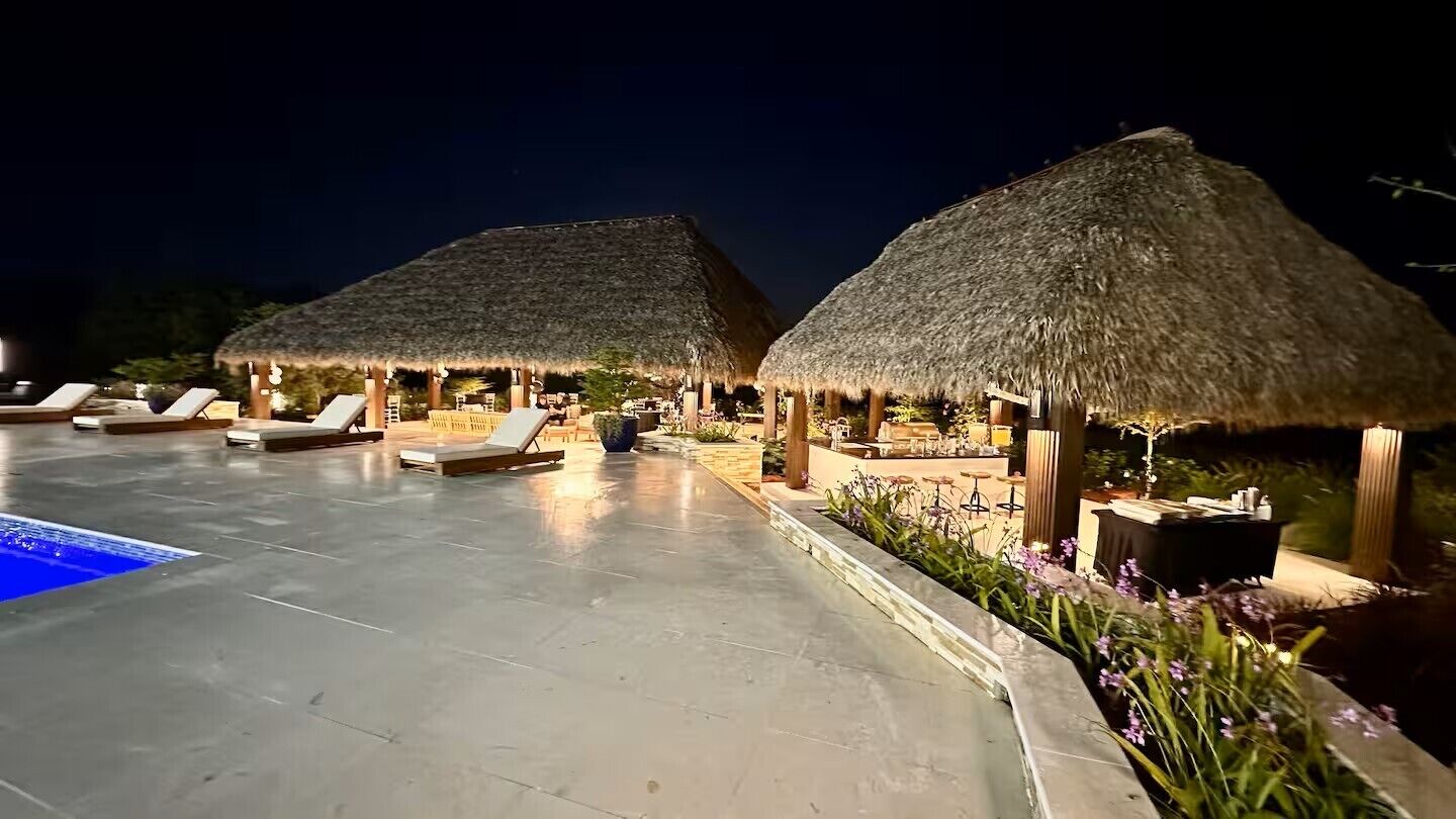 Un área de piscina nocturna con estructuras de techo de paja y sillones de descanso.
