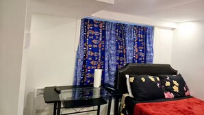 Une petite pièce, de taille modeste, comprenant un lit, une table et une chaise. Il y a une fenêtre avec des rideaux à motifs.