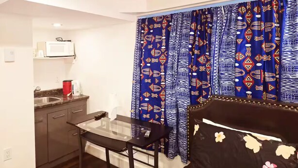 Une chambre d’hôtel compacte comprenant un coin cuisine, une table à manger en verre, un lit avec une literie à motifs floraux et des rideaux à motifs.