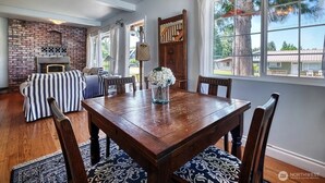 A dining area with a wooden table and chairs, a brick fireplace, striped cushions, and a view of the outdoors through large windows.