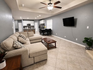 A living room with a sectional sofa, a wooden coffee table, a ceiling fan, and a flat-screen TV mounted on the wall.
