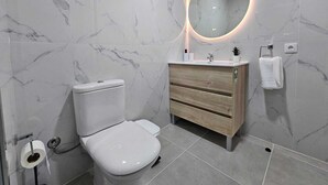 Moderni kylpyhuone, jossa on wc, pesuallas ja pyöreällä valaisimella varustettu peili.
