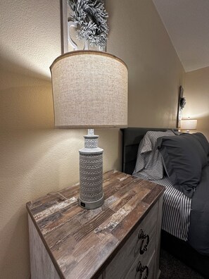 Uma mesa de cabeceira de madeira com uma lâmpada, uma cama com roupa de cama listrada e uma decoração na parede.