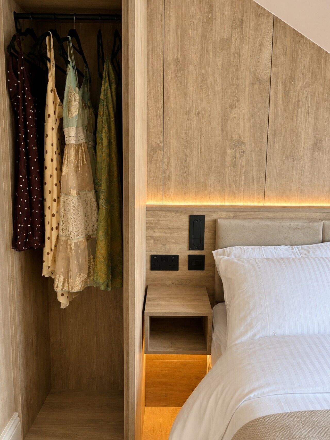 A bedroom with a wooden headboard, a bed with white bedding, a bedside table, and a closet with hanging clothes.
