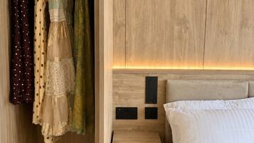 A bedroom with a wooden headboard, a bed with white bedding, a bedside table, and a closet with hanging clothes.