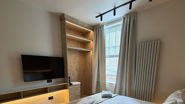 A modern bedroom with a large bed, a TV mounted on the wall, a bedside table, and a window with curtains. - East Parade Collection, Opaz House Flat 2 (North Yorkshire)