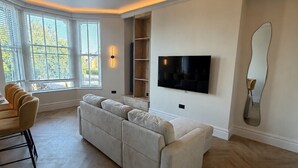 A spacious living room with a large flat-screen TV mounted on the wall, a built-in wooden shelf unit, and a large beige sectional sofa. - East Parade Collection, Opaz House Flat 2 (North Yorkshire)