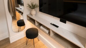 A modern living room with a large flat-screen TV mounted on a wall, a small black ottoman, a white vase with a plant, and a mirror reflecting part of the room. - East Parade Collection, Opaz House Flat 3 (North Yorkshire)