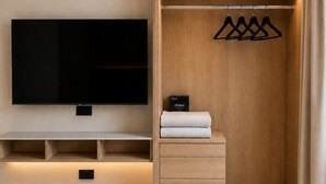 Una habitación moderna con una televisión de pantalla plana montada en la pared, un armario de madera con varios estantes y una barra para colgar ropa, y una mesita de noche con un libro.
