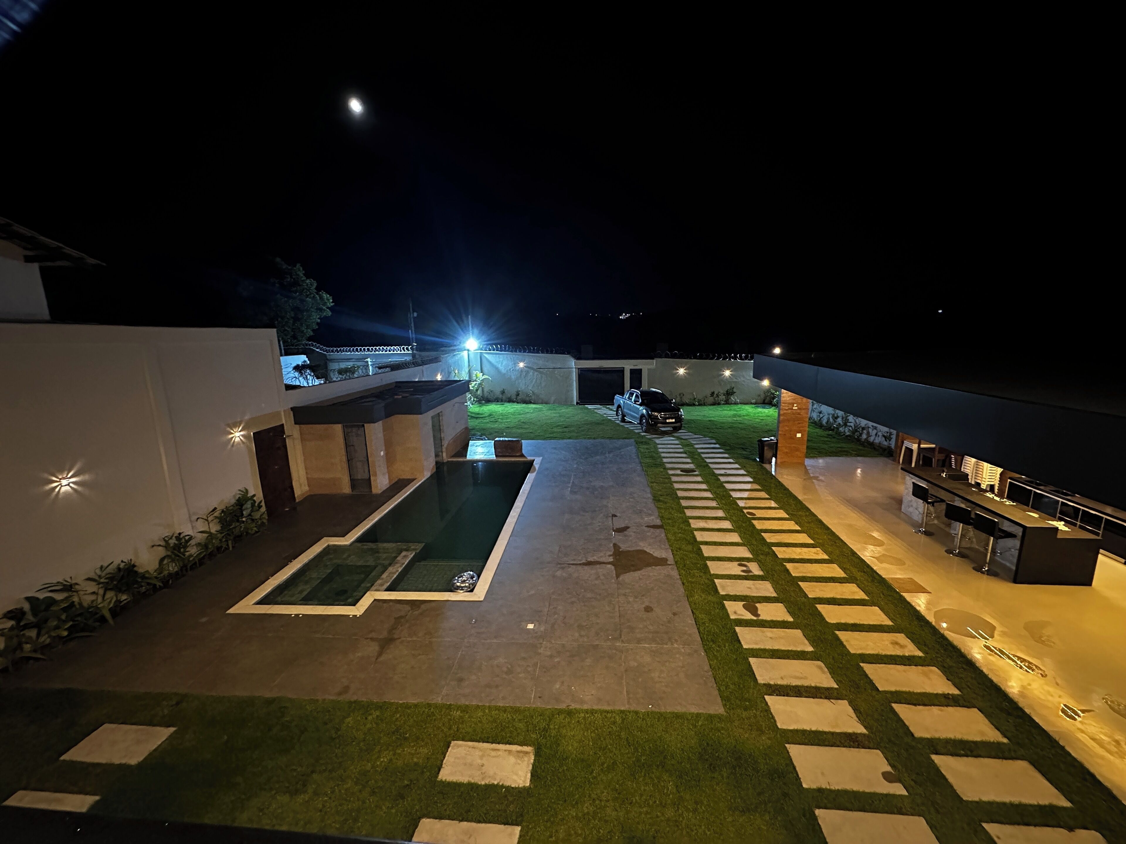 Espaço ao ar livre à noite com piscina, área para churrasco e um caminho que leva a um edifício.