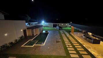 Espaço ao ar livre à noite com piscina, área para churrasco e um caminho que leva a um edifício.