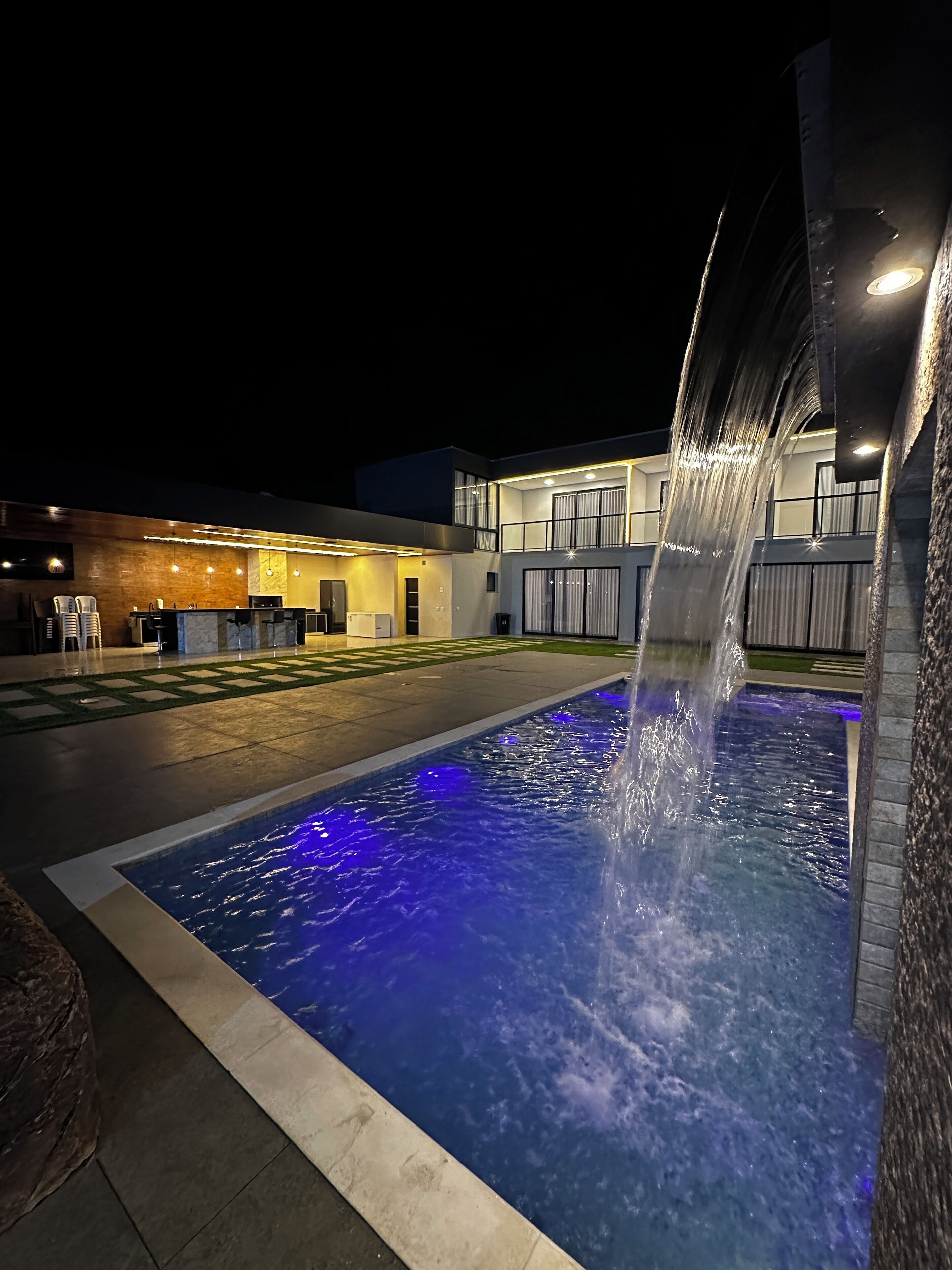 Uma piscina ao ar livre iluminada à noite, com um elemento aquático, um edifício moderno com telhado plano e uma área de estar bem iluminada.