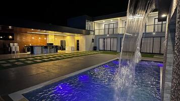 Uma piscina ao ar livre iluminada à noite, com um elemento aquático, um edifício moderno com telhado plano e uma área de estar bem iluminada.