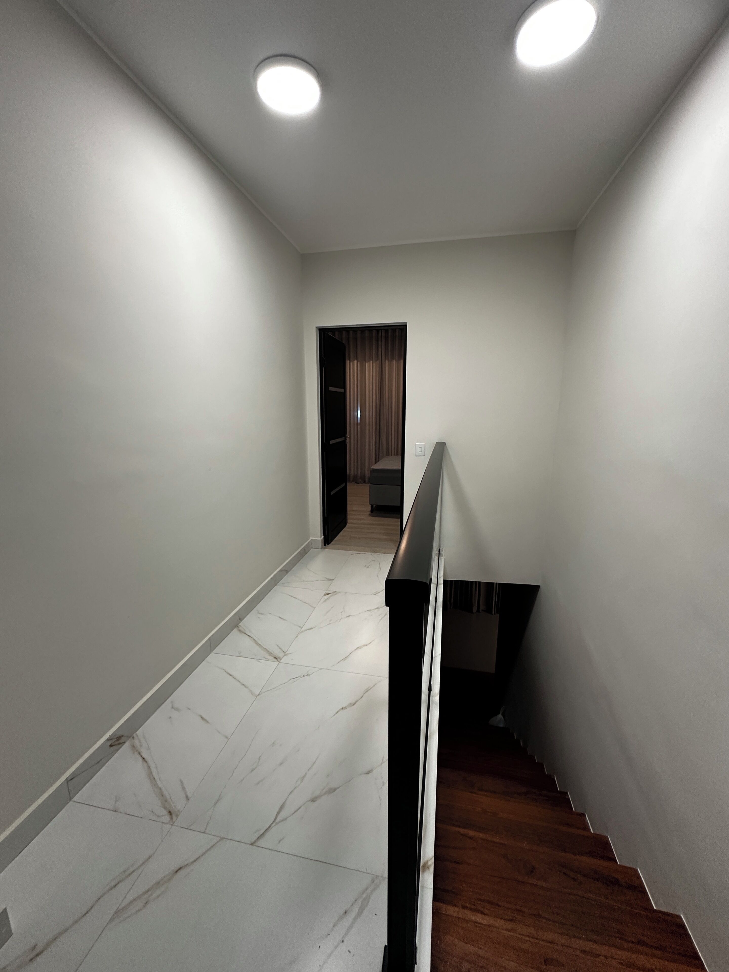 Um corredor com piso de mármore branco, escada de madeira e uma porta que dá para um quarto com cortina.