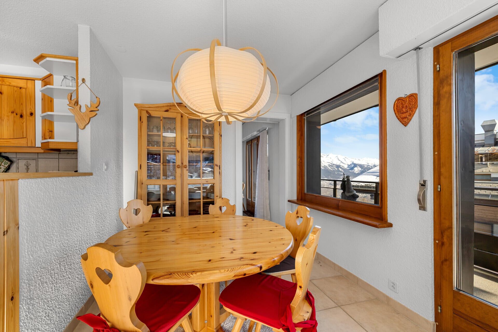Un coin repas avec une table et des chaises en bois, un luminaire suspendu, une fenêtre donnant sur les montagnes et un buffet en bois avec des portes en verre.