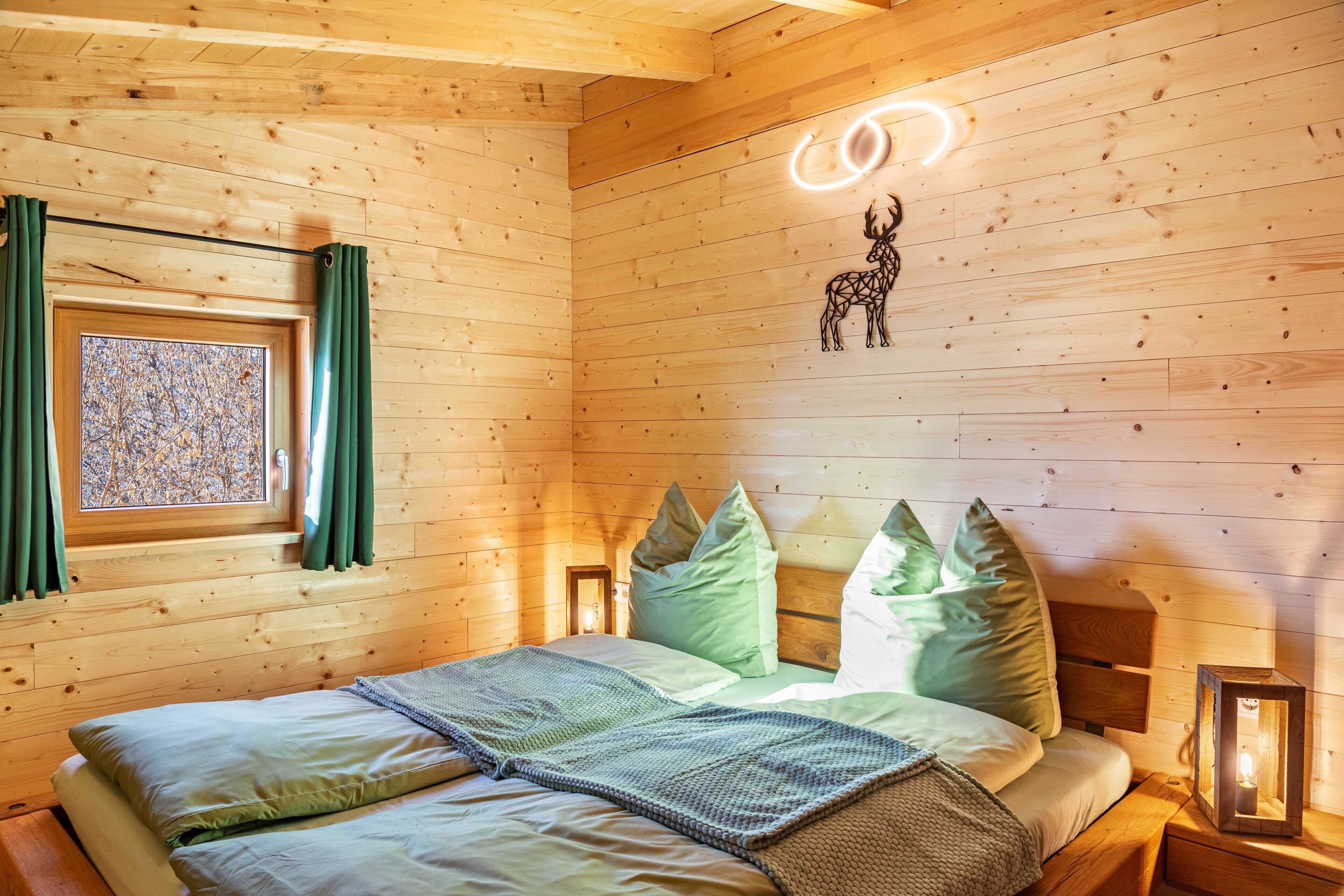 A wooden cabin bedroom with a bed, pillows, a window with curtains, a wall-mounted light, and a decorative animal silhouette.