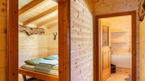 Interior de uma cabana de madeira com uma cama, um cabide e uma porta para outro cômodo.