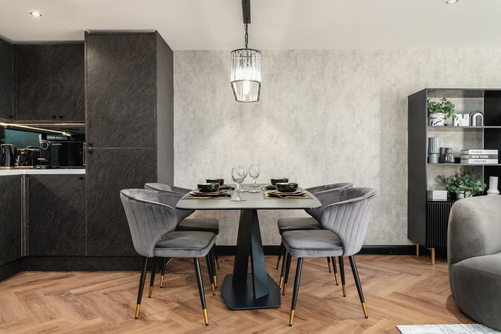 Un comedor moderno con suelo de madera en espiga oscura, una mesa central con cuatro sillas tapizadas en gris y una lámpara colgante.
