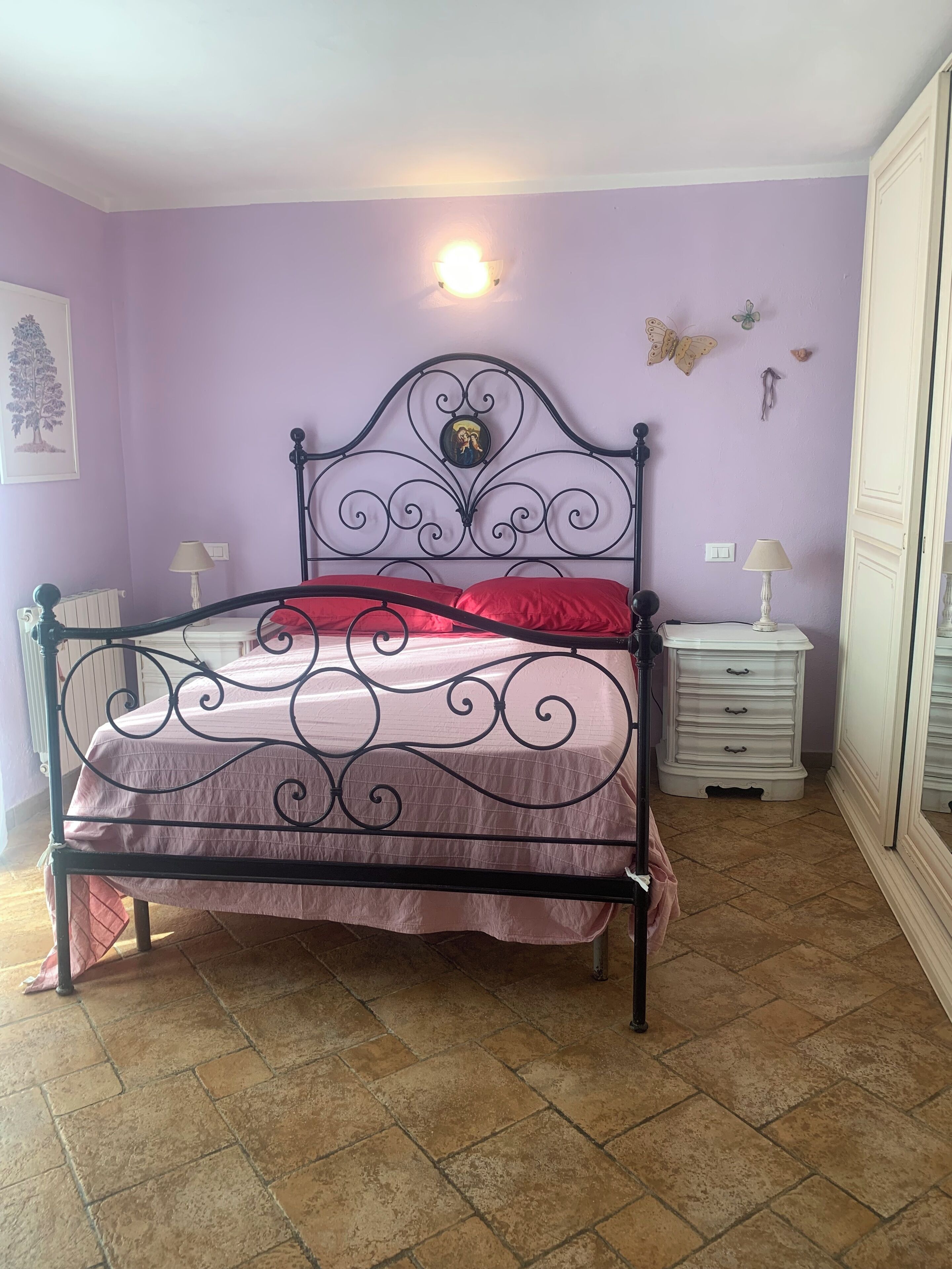 A bed with a metal frame and a headboard with a decorative design, a red pillow, and a white nightstand with a lamp.