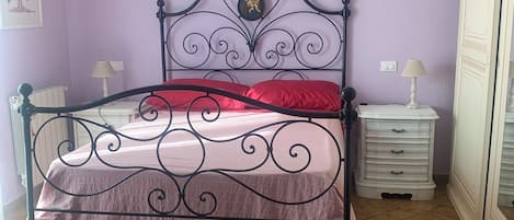 A bed with a metal frame and a headboard with a decorative design, a red pillow, and a white nightstand with a lamp.