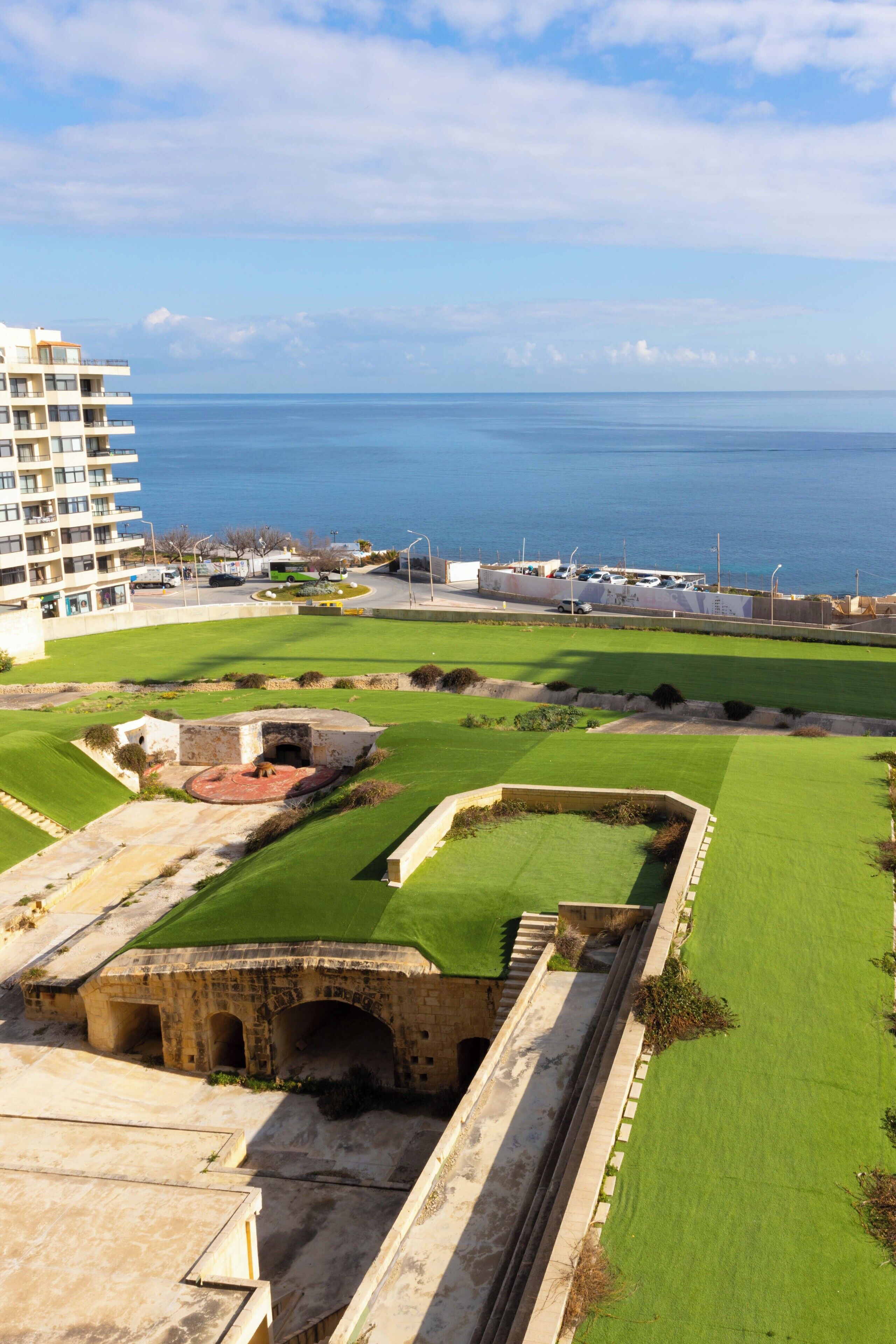 A coastal view with a grassy area, a stone structure, and a modern building in the background.
