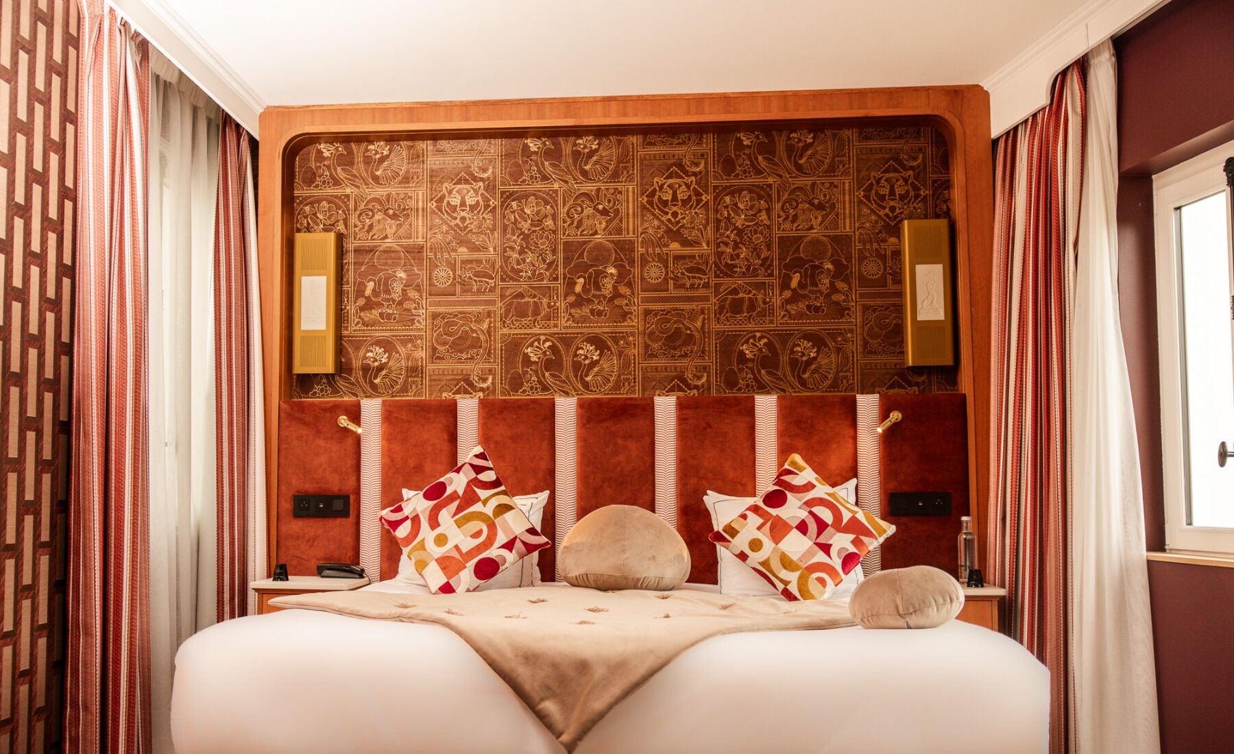 Quarto de hotel com cabeceira estampada, cama com várias almofadas, mesa de cabeceira com abajur e cortinas.