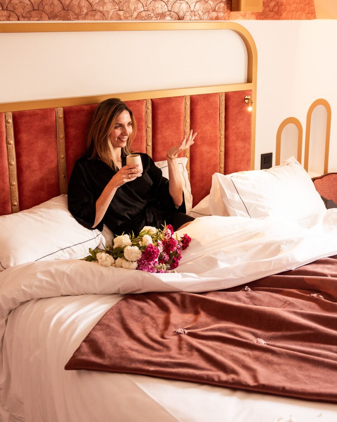 A bed with a headboard, a bouquet of flowers, and a person sitting on the bed.