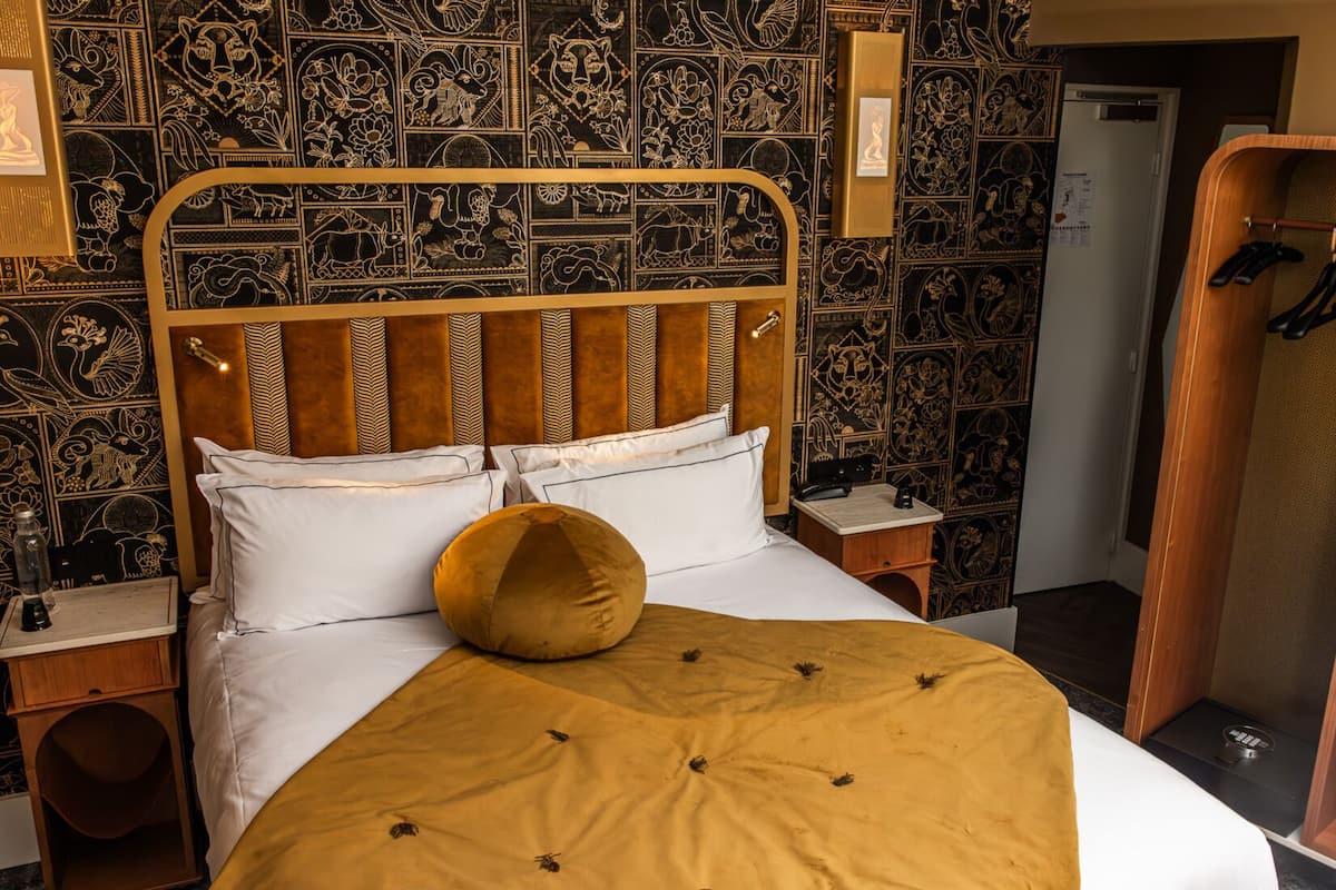 A bed with a headboard, white bedding, and a mustard-colored pillow. There are two bedside tables with lamps. The room has a patterned wallpaper and a wooden wardrobe.