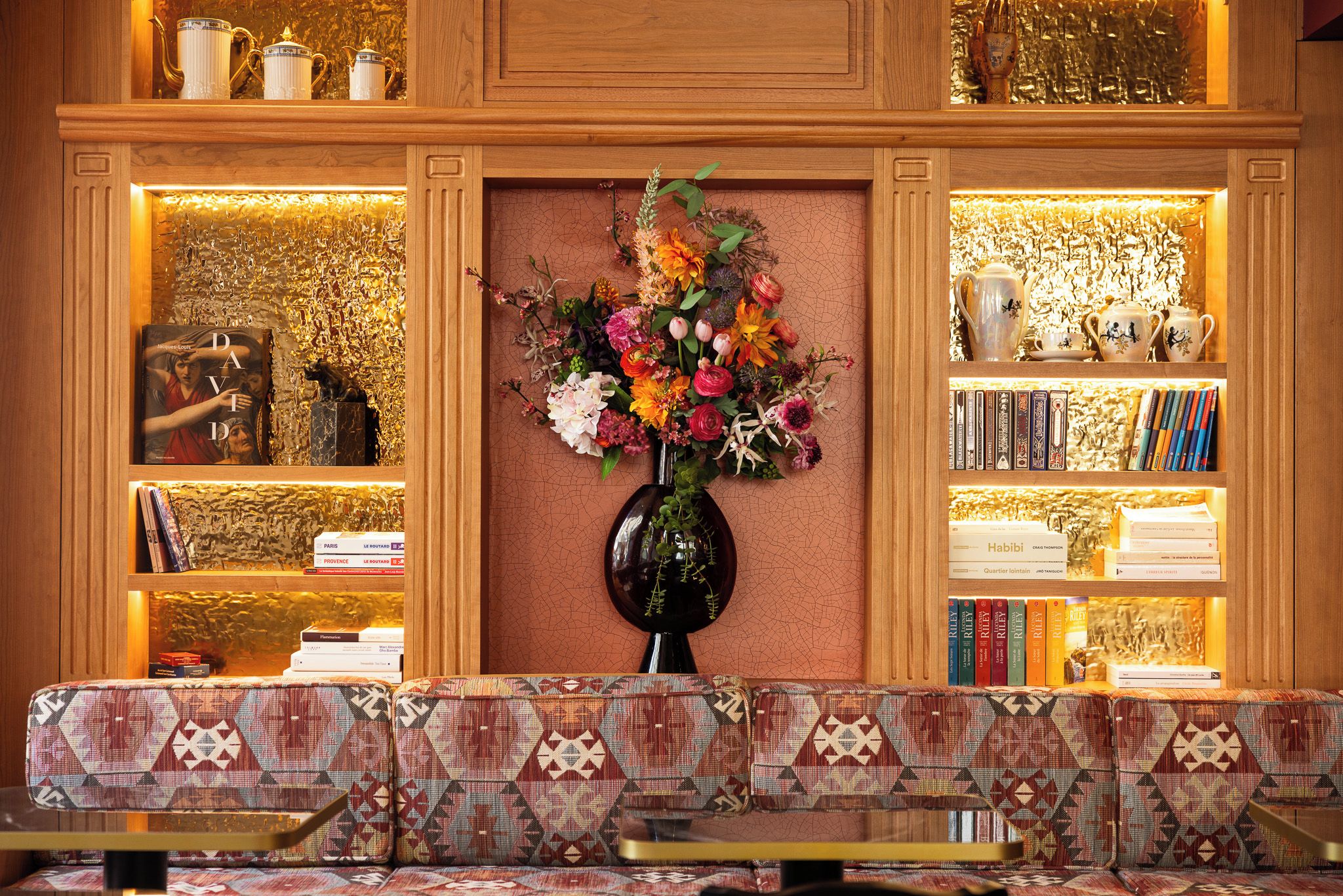 Un espace d’accueil agrémenté d’un canapé à motifs, d’un vase de fleurs et d’étagères où sont disposés des livres et des objets décoratifs.