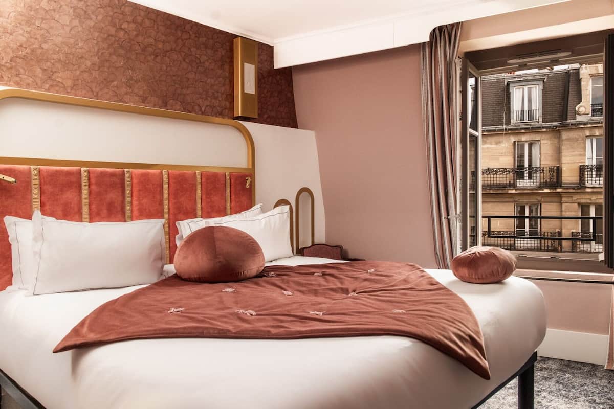 A hotel room with a bed, a headboard, a window with a view of buildings, and a curtain.