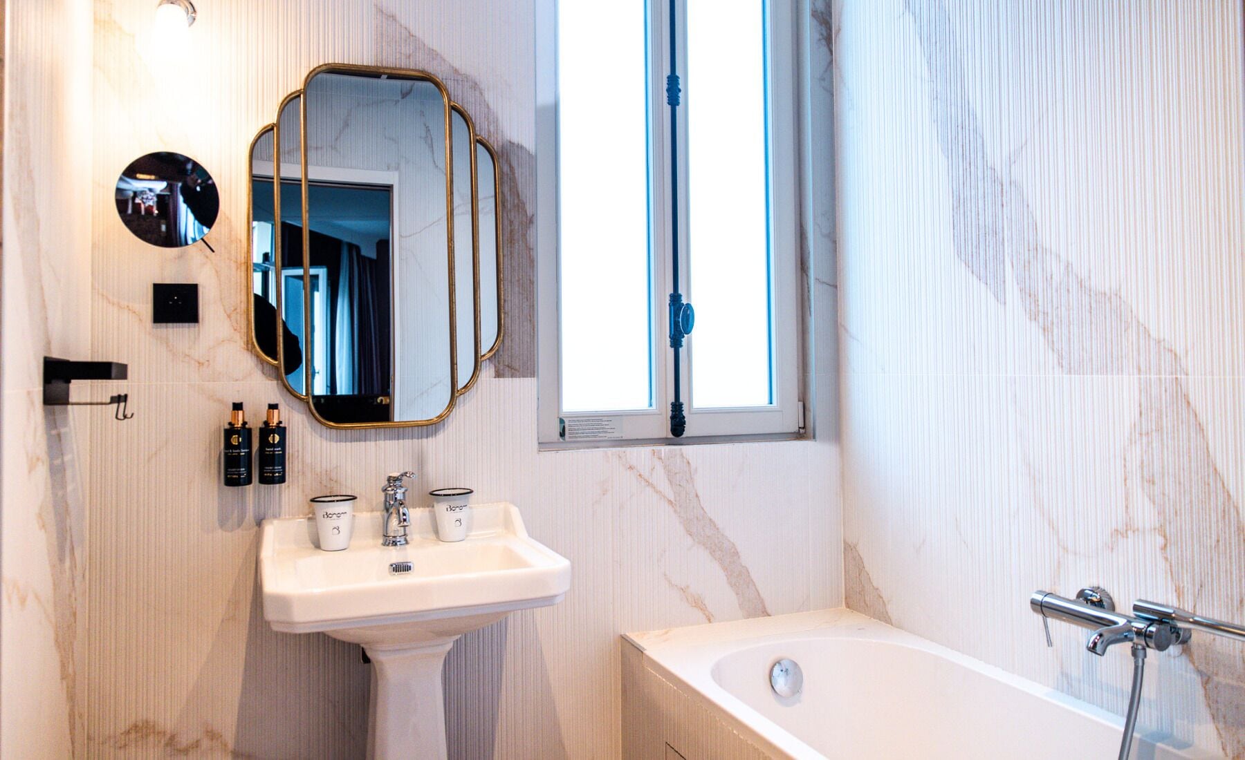 Ein Badezimmer mit Marmorwand, einem Waschbecken mit zwei Flaschen, einem Spiegel und einem Fenster.