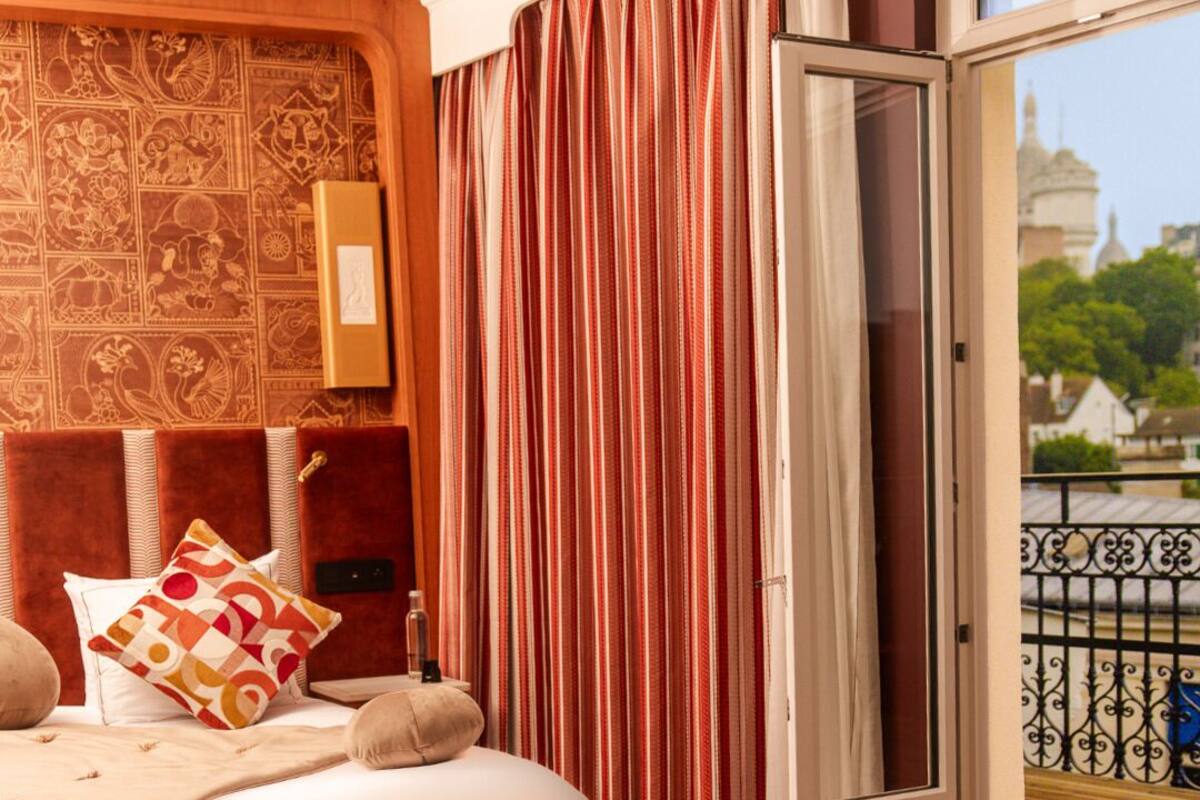 A hotel room with a bed, a wooden headboard, a patterned wall, a window with curtains, and a balcony view.