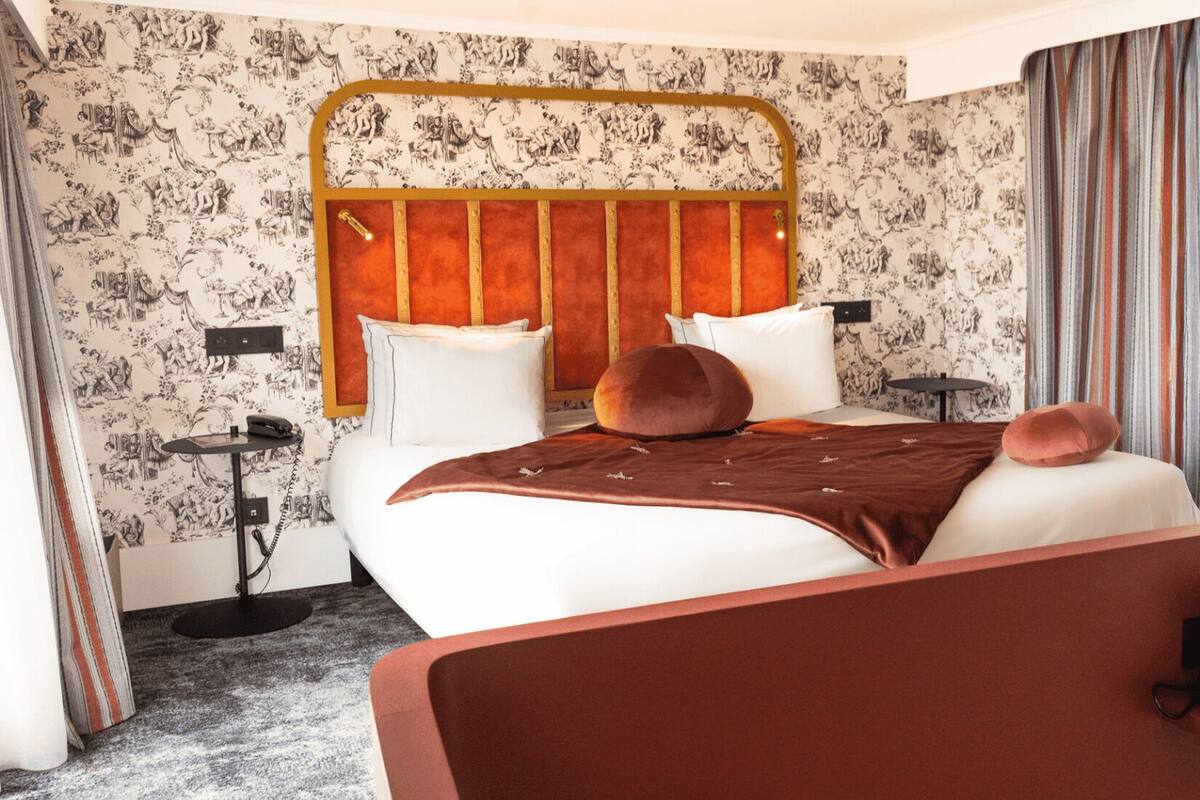 A hotel room with a large bed, a headboard with vertical lines, a bedside table, a telephone, and a window with curtains.