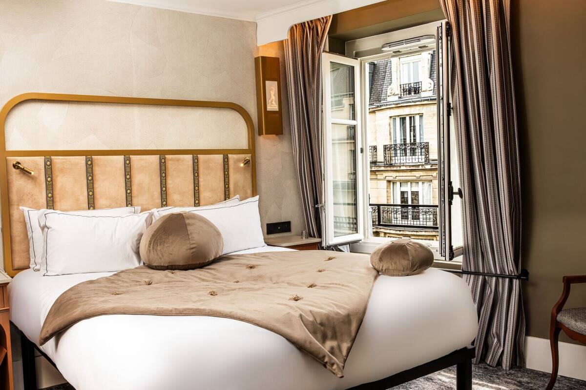 A hotel room with a bed, a bedside table, a chair, a window with curtains, and a view of a building.
