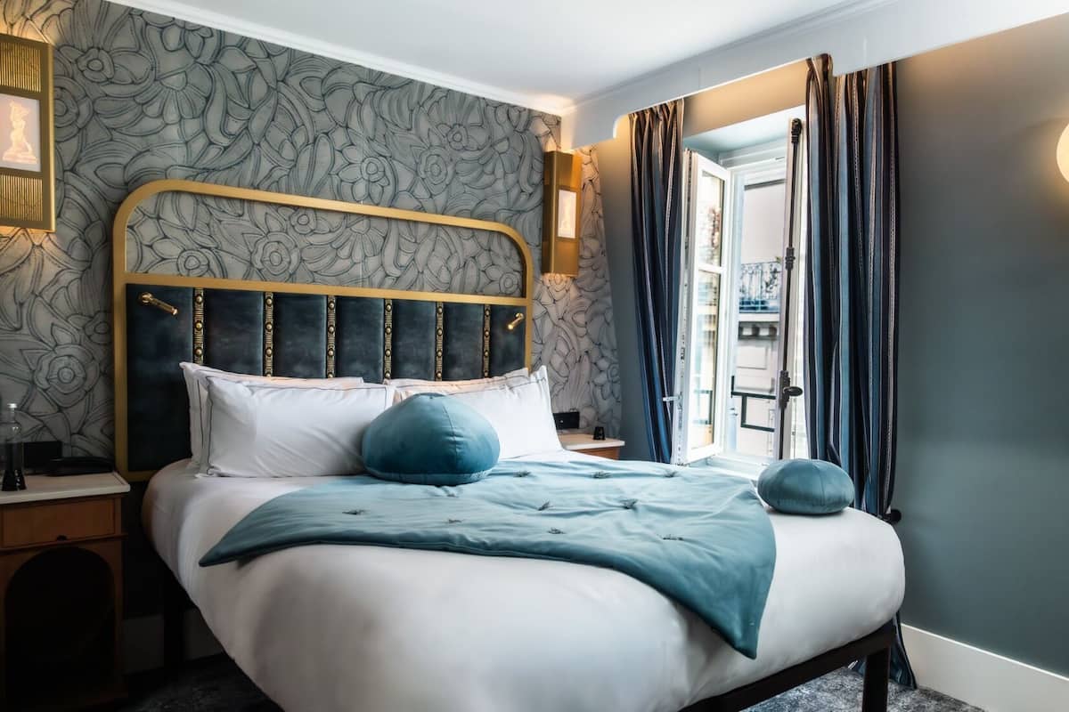 A neatly made bed with a headboard, a bedside table, and a nightstand. There is a window with curtains, a wall-mounted light, and a door leading to another room.