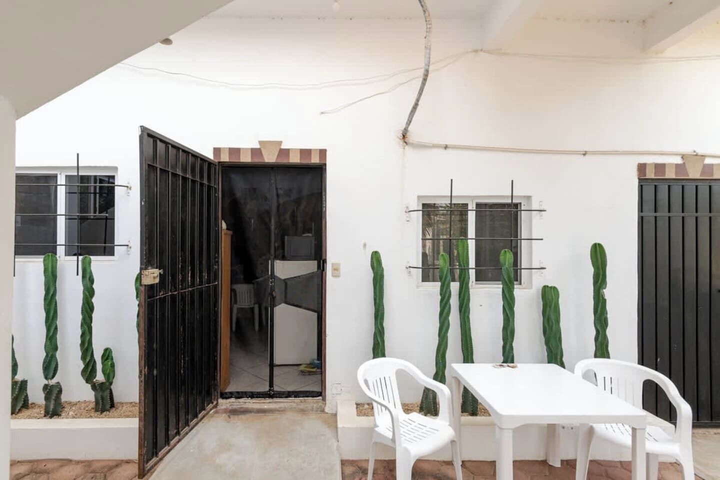 Un espace patio avec une table et des chaises blanches, entouré de cactus, et un portail noir donnant accès à une pièce.