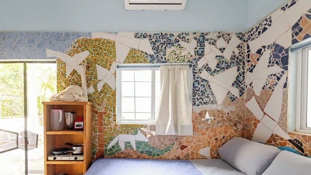 A bedroom with a mosaic wall, a window with curtains, a bed with pillows, and a wooden shelf with kitchen items. - Steps from the beach - easy checkin (Brisas de Zicatela)