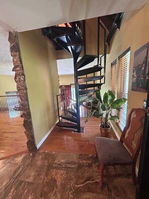 Una escalera con diseño espiral que conduce a un nivel superior, una planta en maceta y una silla de madera en una habitación con suelo de madera y un muro de piedra decorativo.