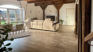 A spacious living room with a high ceiling and wooden beams, a large sofa, a TV, and a view of the outdoors through a glass door. - Vacation on the farm for 6 pers. (Arlewatt)