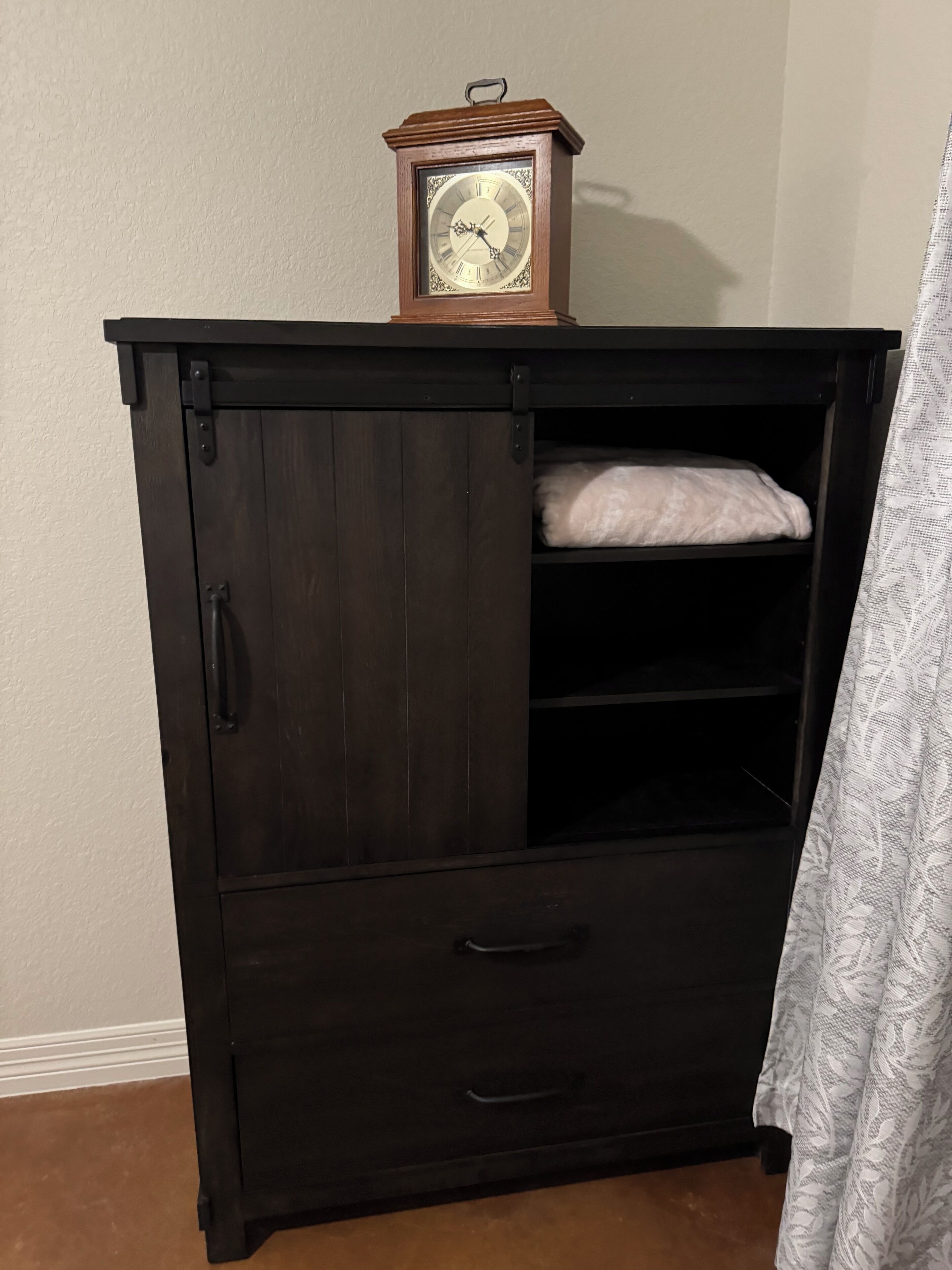 A dark wooden cabinet with a clock on top, a shelf with a folded white cloth, and a drawer with a handle.