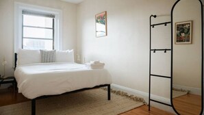 Ein Schlafzimmer mit Bett, Fenster, Nachttisch, Spiegel und einem Wandregal.