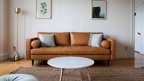 Ein Wohnzimmer mit einem braunen Ledersofa, zwei gerahmten Bildern an der Wand, einer Stehlampe und einem runden Couchtisch.