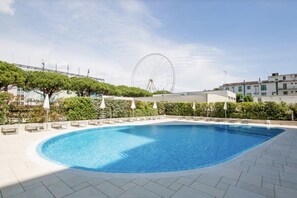 Ein Pool mit geschwungener Kante, umgeben von einer gefliesten Terrasse. Im Hintergrund sieht man ein Riesenrad und eine Reihe von Gebäuden.