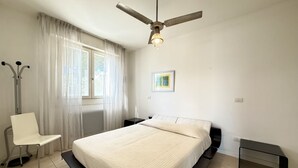Un dormitorio con cama, una silla, un escritorio, un ventilador de techo y una ventana con cortinas.