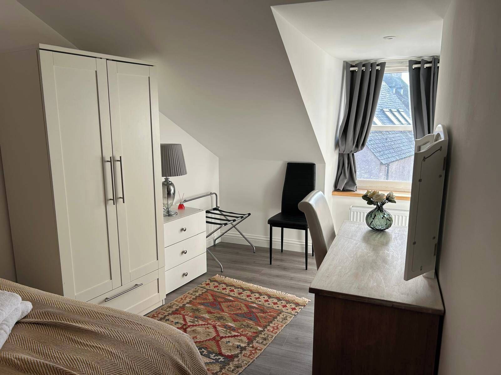 A bedroom with a bed, a desk with a lamp, a chair, a wardrobe, and a window with curtains.